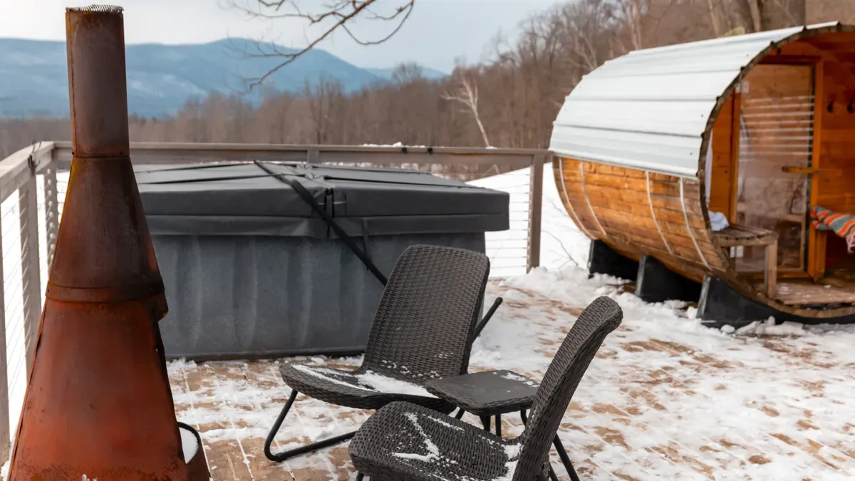 A snow-covered wooden deck with a covered hot tub, outdoor chairs, and a barrel-style wooden sauna in a winter mountain setting.